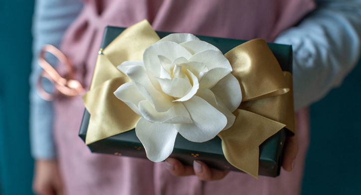 A crafter handing the viewer a gift with a beauiful flower bow.